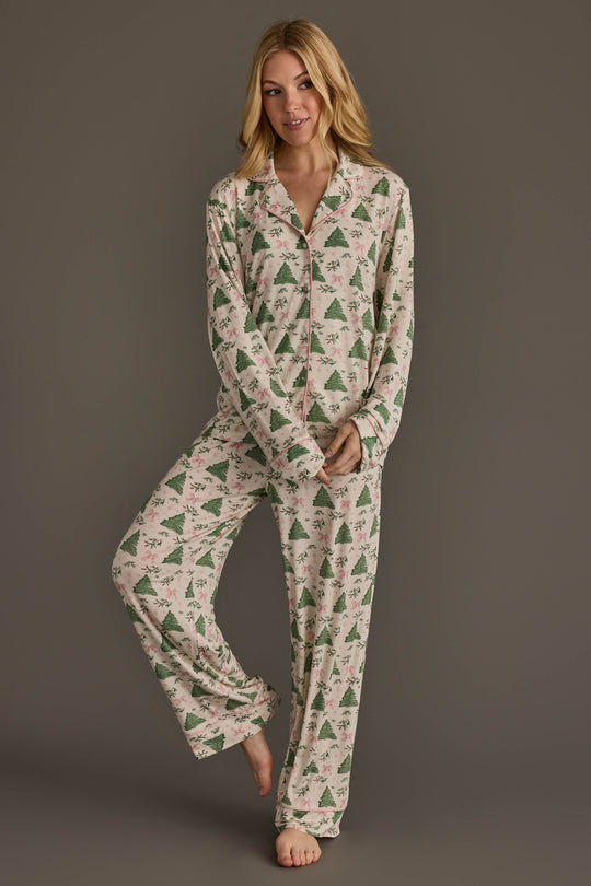 A woman wears a Panache Apparel Collection Trees & Bows Jersey Pajama Set consisting of a long-sleeved button-down top and matching pants patterned with green pine trees and pink bows.