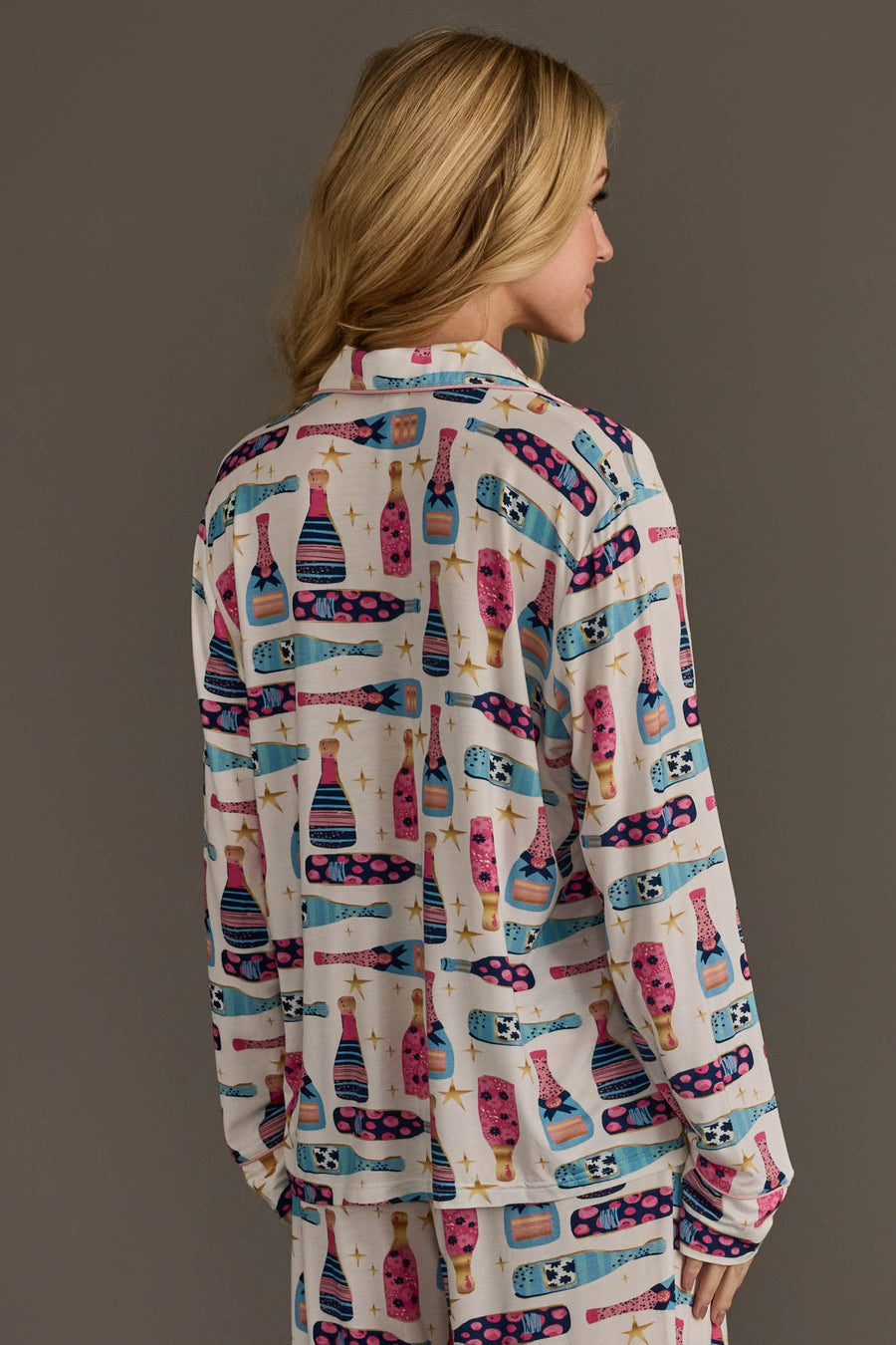 A person wears a Panache Apparel Collection Champagne Bottles Jersey Pajama Set consisting of long-sleeved button-down pajamas decorated with a pattern of colorful bottles and gold stars.