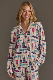 A smiling woman wears a Panache Apparel Collection Champagne Bottles Jersey Pajama Set decorated with colorful champagne bottles and gold stars.
