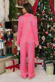 A woman stands with her back to the camera wearing a Red & Pink Striped Satin Pajama Set featuring a long-sleeved button-down top and matching pants with red and pink vertical stripes and white piping.
