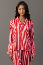 A person wears a Red & Pink Striped Satin Pajama Set consisting of a long-sleeved, button-down top and matching pants in a red and pink striped pattern with white piping.