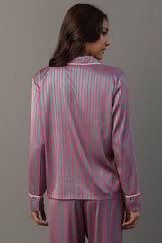 A woman wears a Pink & Green Striped Satin Pajama Set consisting of a long-sleeved button-up top and matching pants with pink and green vertical stripes and white piping.