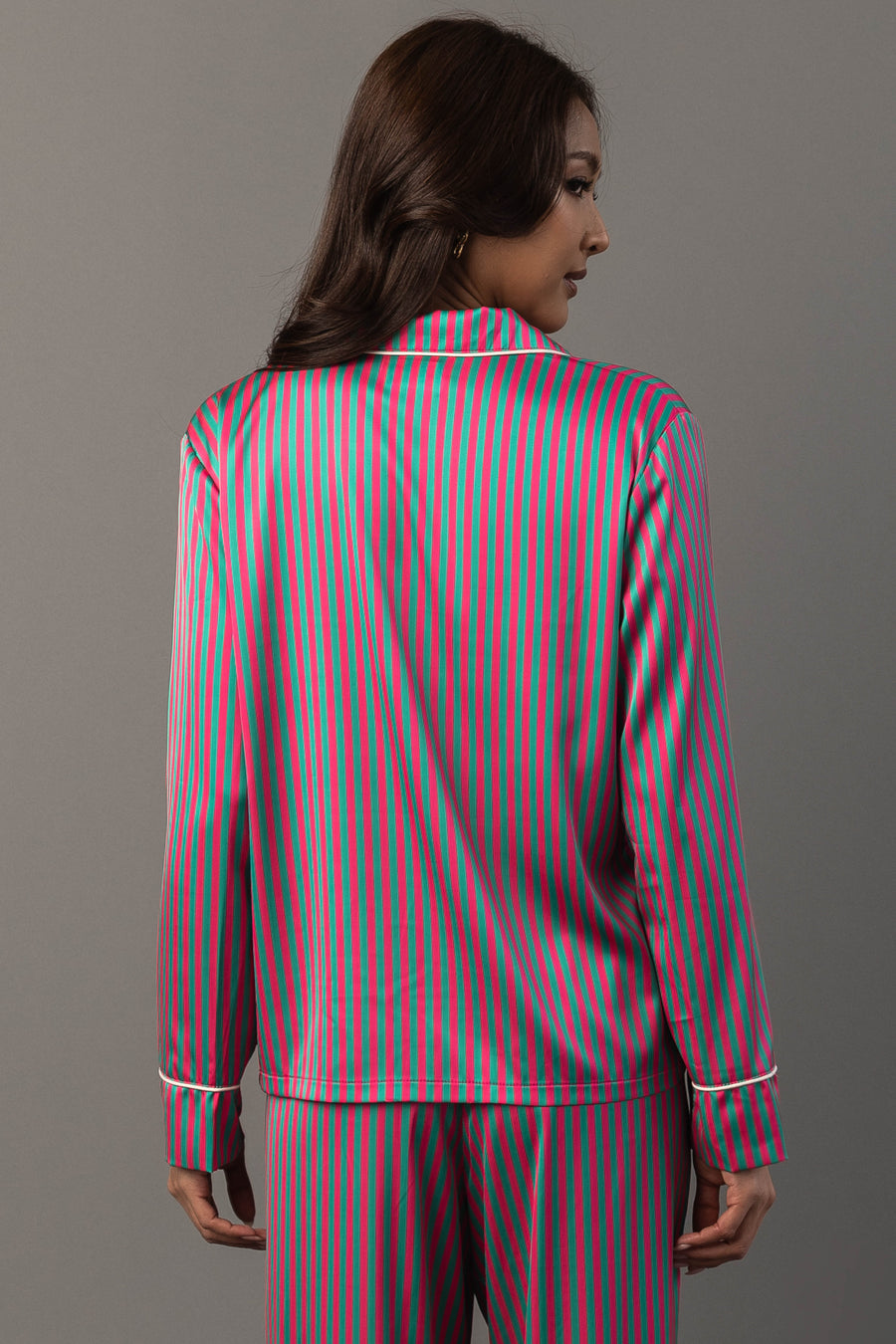 A woman wears a Pink & Green Striped Satin Pajama Set consisting of a long-sleeved button-up top and matching pants with pink and green vertical stripes and white piping.