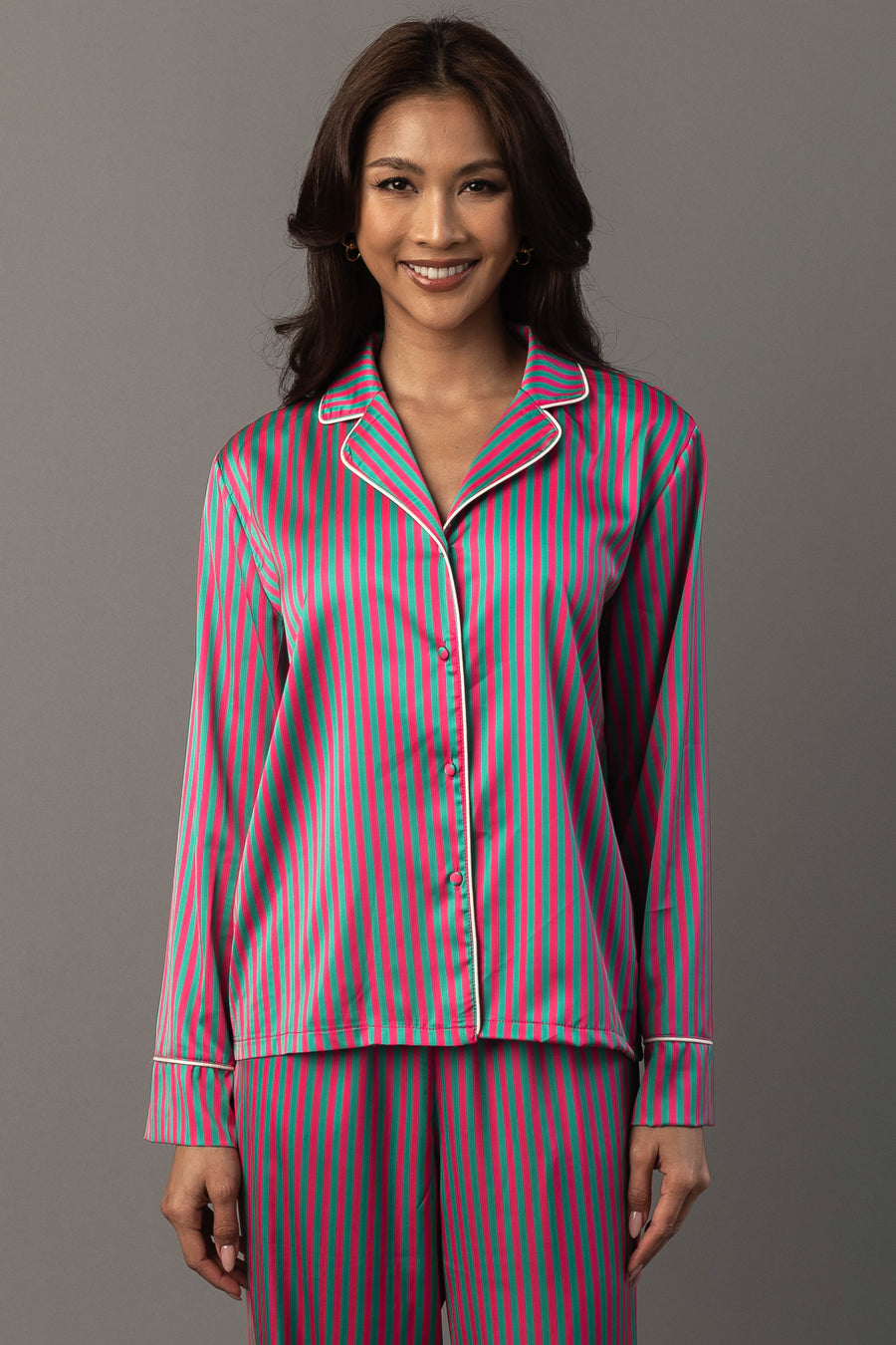 A smiling woman wears a Pink & Green Striped Satin Pajama Set featuring a long-sleeved button-down top and matching pants with pink and green vertical stripes and white piping.
