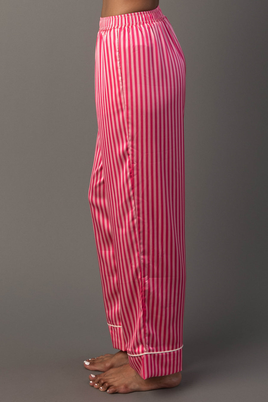 Profile view of Bright Pink & Light Pink Striped Satin Pajama Set pants featuring vertical bright and light pink stripes, an elastic waistband, and white trim at the cuffs.