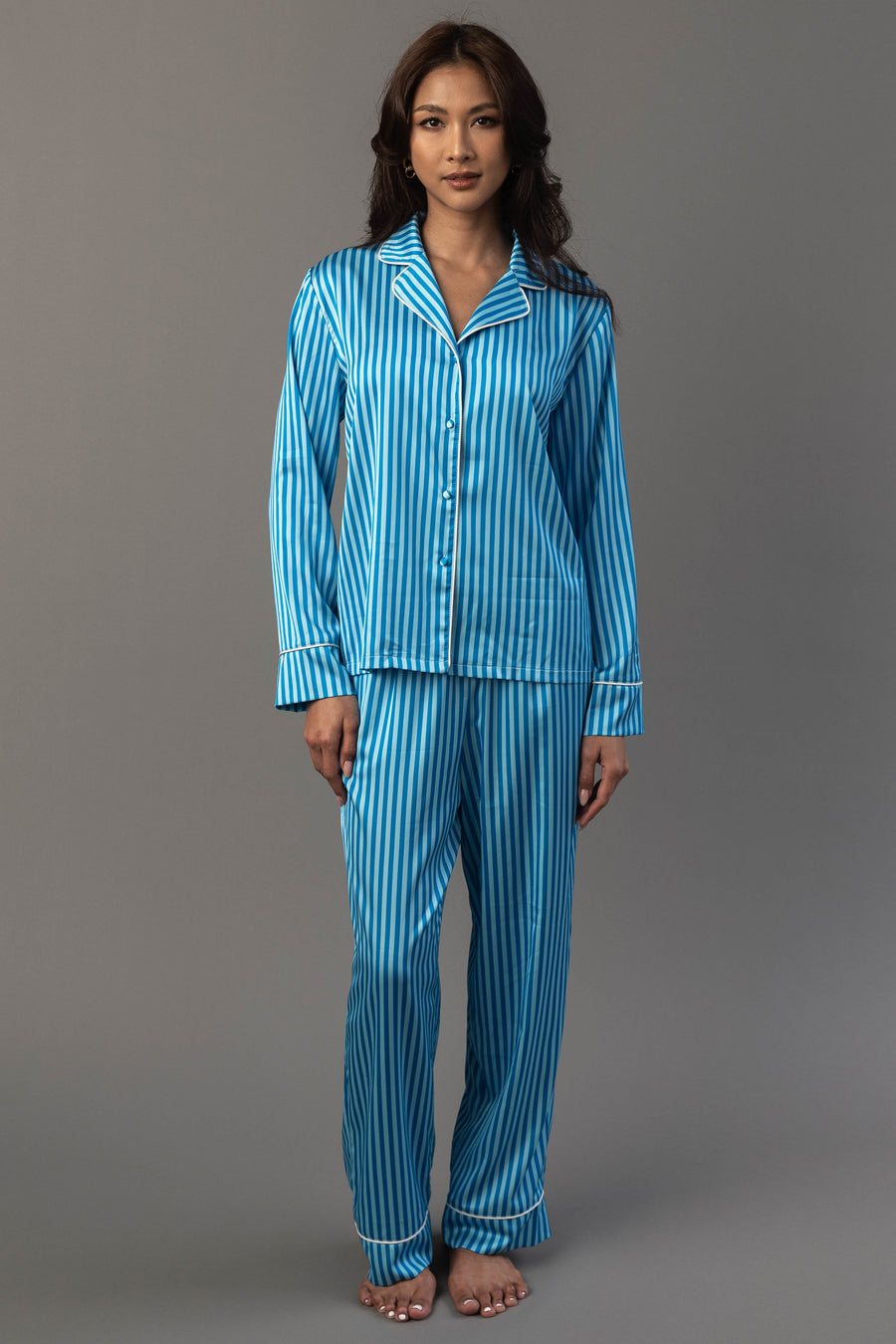 A person wearing a Blue & Light Blue Striped Satin Pajama Set, featuring a long-sleeved button-down top and matching pants in blue and light blue vertical stripes with white trim.