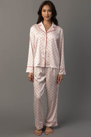 A woman wears an Ivory & Red Ho Ho Ho Satin Pajama Set, featuring a long-sleeved button-up ivory top and pants with a repeating red Ho Ho Ho print and red piping.