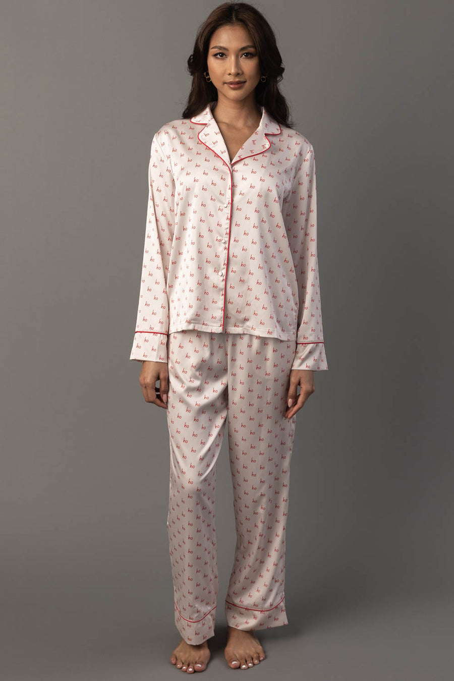 A woman wears an Ivory & Red Ho Ho Ho Satin Pajama Set, featuring a long-sleeved button-up ivory top and pants with a repeating red Ho Ho Ho print and red piping.