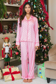A woman stands in front of a Christmas tree wearing a Light Pink & Red Ho Ho Ho Satin Pajama Set, featuring a button-down collared shirt and matching pants in light pink with a repeating red ho ho ho pattern.