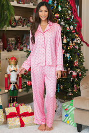 A person wearing a Light Pink & Red Ho Ho Ho Satin Pajama Set with a repeating ho ho ho pattern and red piping, standing in front of a decorated Christmas tree.