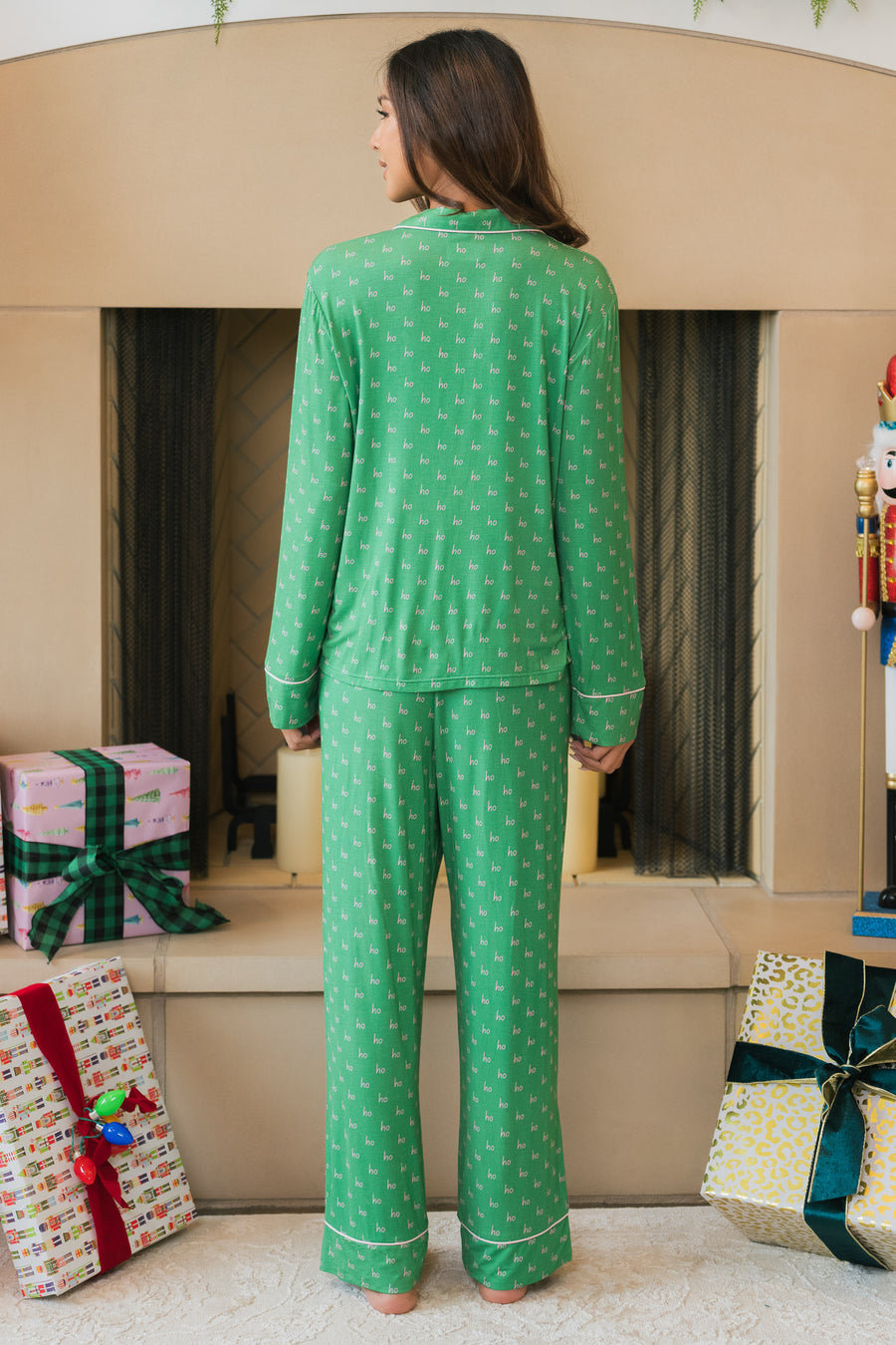 A person stands with their back to the camera wearing a Green & Light Pink Ho Ho Ho Jersey Pajama Set featuring a repeating ho ho ho pattern and light pink trim.