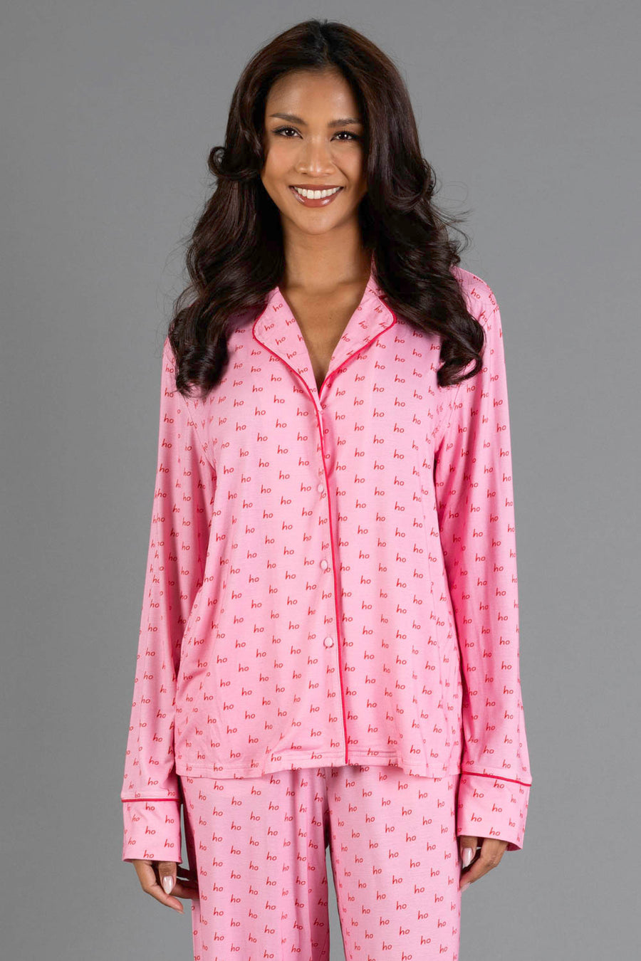 A woman wearing a Light Pink & Red Ho Ho Ho Jersey Pajama Set consisting of a long-sleeved top and pants featuring an all-over red ho ho ho print.