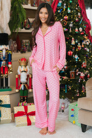 A smiling woman stands before a Christmas tree and nutcracker figurines wearing a light pink and red ho ho ho Light Pink & Red Ho Ho Ho Jersey Pajama Set from Panache Apparel Collection.