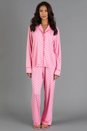 A woman wearing a Light Pink & Red Ho Ho Ho Jersey Pajama Set, featuring a light pink jersey pajama set with a repeating small red ho ho ho print and red piping trim on the long-sleeved button-down top and matching pants.