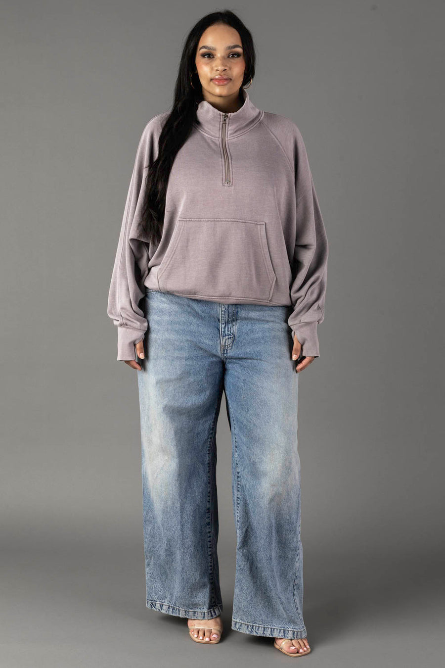 A woman with long dark hair and a relaxed expression stands facing forward against a plain gray background, wearing the Panache Apparel Collection Gray Quarter Zip Sweatshirt, light blue wide-leg jeans, and open-toed heels.