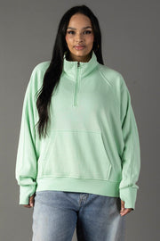 A woman with long dark hair and a neutral expression wears the Panache Apparel Collection Mint Quarter Zip Sweatshirt, featuring a cropped fit, funnel neck, front pocket, and light green color, paired with blue jeans against a gray background.