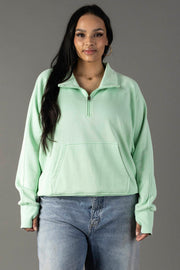 A woman with long dark hair wears the Panache Apparel Collection Mint Quarter Zip Sweatshirt, featuring long sleeves and thumbholes, paired with light blue jeans, standing against a plain gray background and smiling softly at the camera.