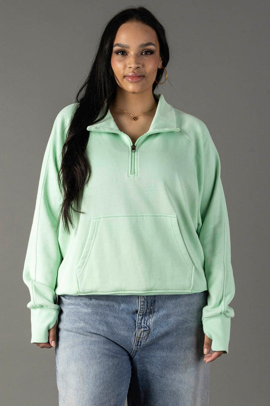 A woman with long dark hair wears the Panache Apparel Collection Mint Quarter Zip Sweatshirt, featuring long sleeves and thumbholes, paired with light blue jeans, standing against a plain gray background and smiling softly at the camera.