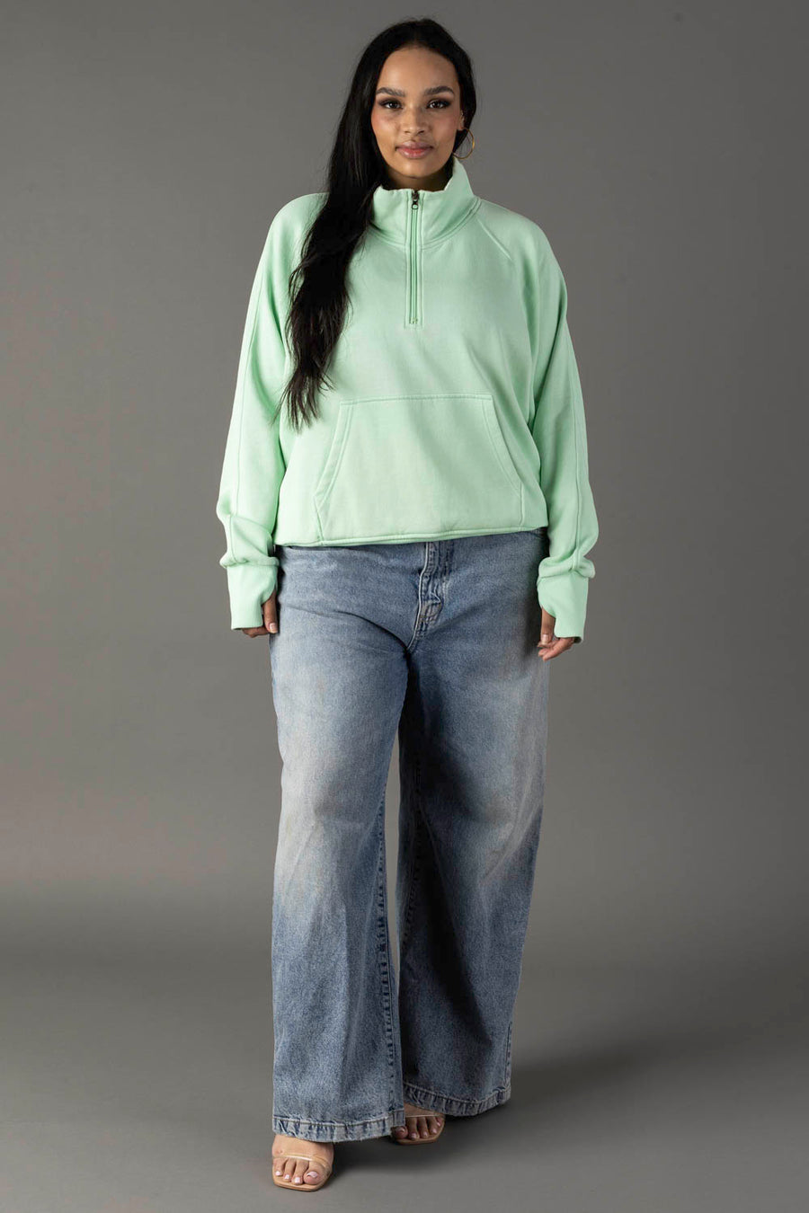 A woman with long dark hair stands facing forward against a gray background, wearing the Panache Apparel Collection Mint Quarter Zip Sweatshirt with thumbholes and wide-leg blue jeans. She is also wearing open-toed shoes.