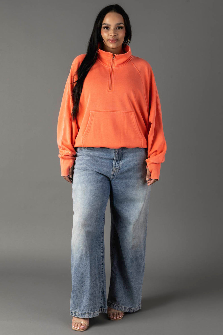 A woman with long dark hair and a confident look models the Panache Apparel Collection's Coral Quarter Zip Sweatshirt, featuring a front pocket, paired with wide-leg blue jeans and open-toe heels against a gray background.