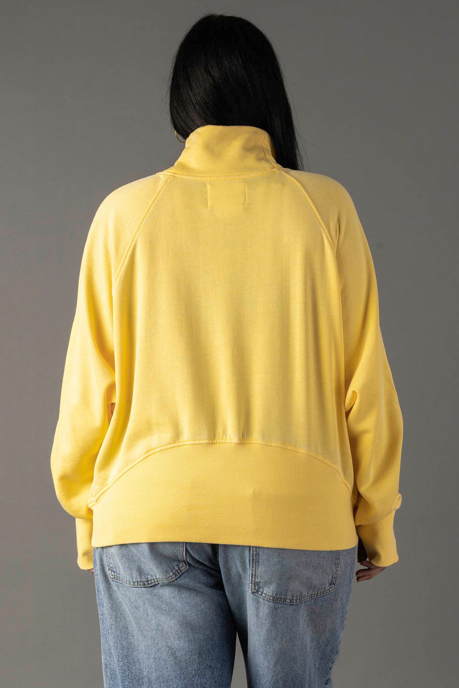 A person with long dark hair is seen from the back, wearing a Panache Apparel Collection Yellow Quarter Zip Sweatshirt and light blue jeans against a plain gray background.