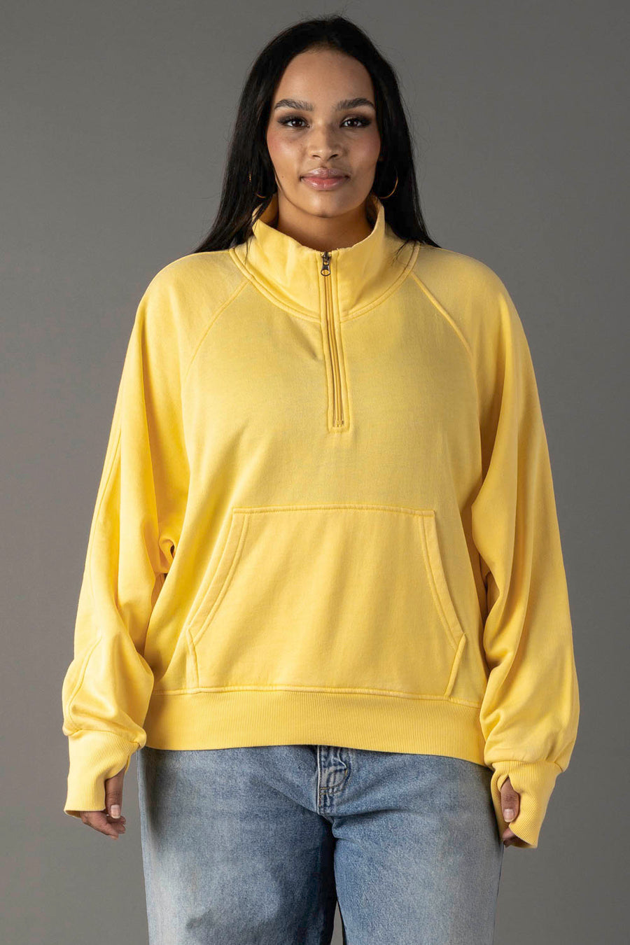 A woman faces forward in a loose-fitting Panache Apparel Collection Yellow Quarter Zip Sweatshirt with a kangaroo pocket, paired with light blue jeans. The background is plain gray.