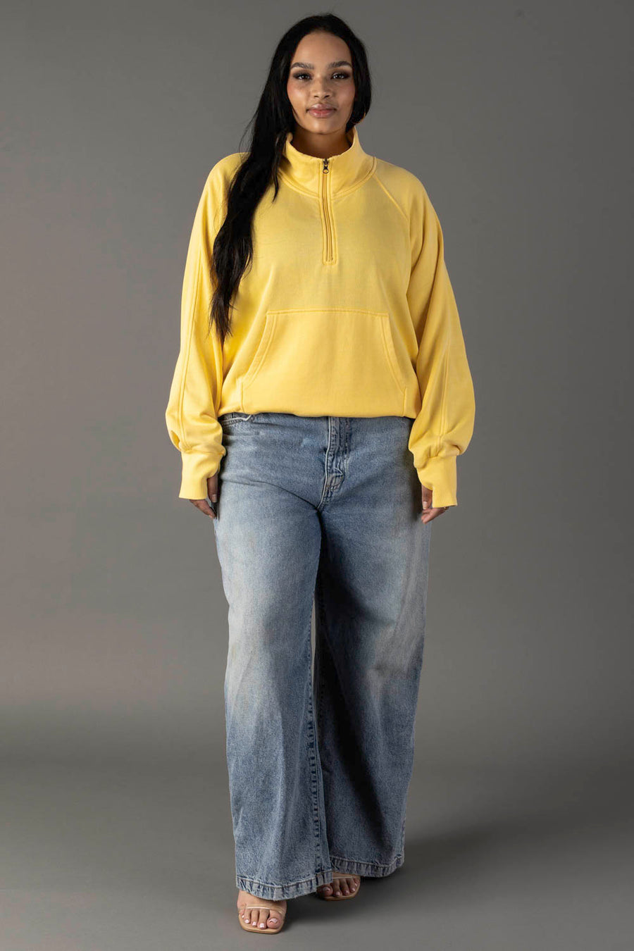 A woman with long dark hair stands against a gray background in a Panache Apparel Collection Yellow Quarter Zip Sweatshirt, wide-leg blue jeans, and open-toe sandals.