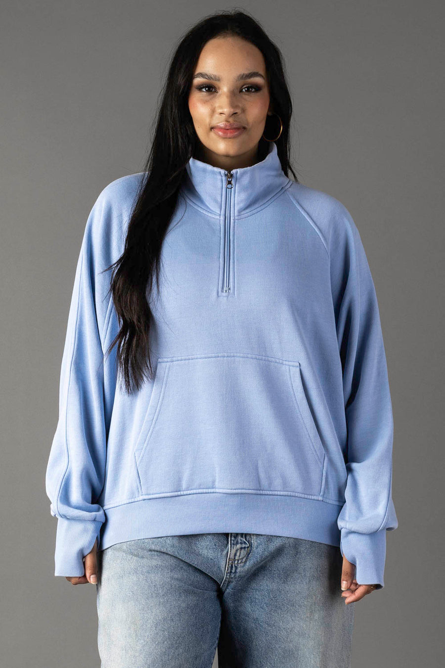 A woman with long dark hair models the Panache Apparel Collection Light Blue Quarter Zip Sweatshirt, paired with light blue jeans, standing against a plain gray background and smiling gently at the camera.