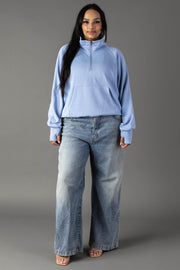 A woman with long dark hair, facing forward with a slight smile, wears the Panache Apparel Collection Light Blue Quarter Zip Sweatshirt, paired with loose blue jeans and open-toe heels against a gray background.