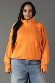 A woman with long dark hair wears the Orange Quarter Zip Sweatshirt by Panache Apparel Collection and light blue jeans, standing against a plain gray background and smiling slightly.