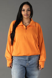 A woman with long dark hair models the Panache Apparel Collection Orange Quarter Zip Sweatshirt and light blue jeans, standing against a gray background and smiling slightly at the camera.