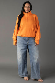 Against a gray background, a woman with long dark hair and a relaxed look models the Panache Apparel Collection Orange Quarter Zip Sweatshirt with front pocket, paired with loose blue jeans and open-toed sandals.