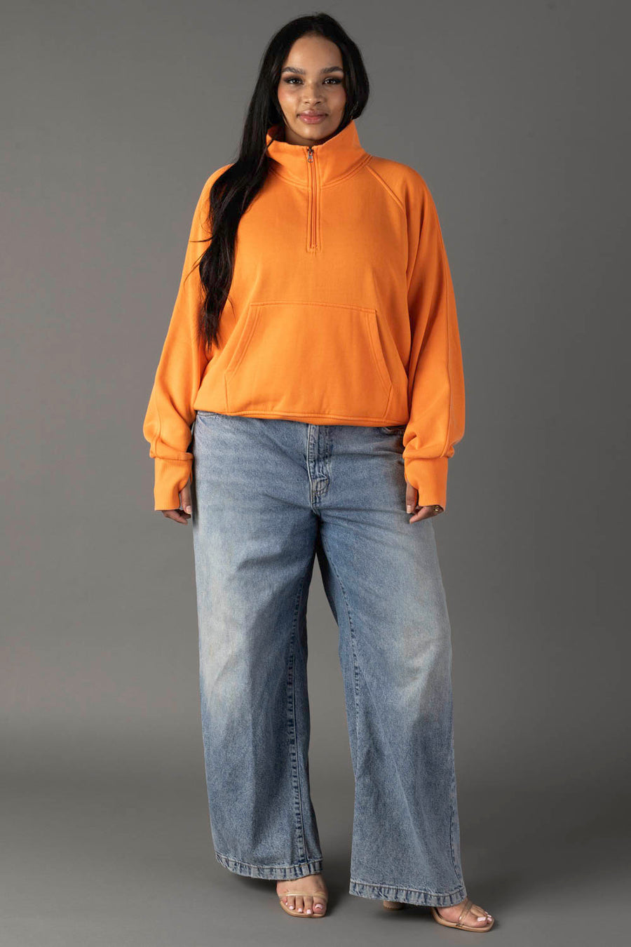 Against a gray background, a woman with long dark hair and a relaxed look models the Panache Apparel Collection Orange Quarter Zip Sweatshirt with front pocket, paired with loose blue jeans and open-toed sandals.