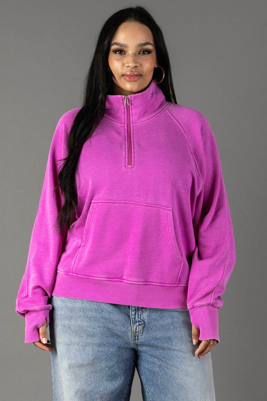 A woman with long dark hair smiles gently at the camera, wearing the Purple Quarter Zip Sweatshirt from Panache Apparel Collection and light blue jeans, standing against a plain gray background.