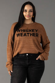 A woman with long dark hair models the Brown Whiskey Weather Lightweight Sweater from Panache Apparel Collection, styled with black jeans. She stands against a gray background, smiling with one hand in her pocket.