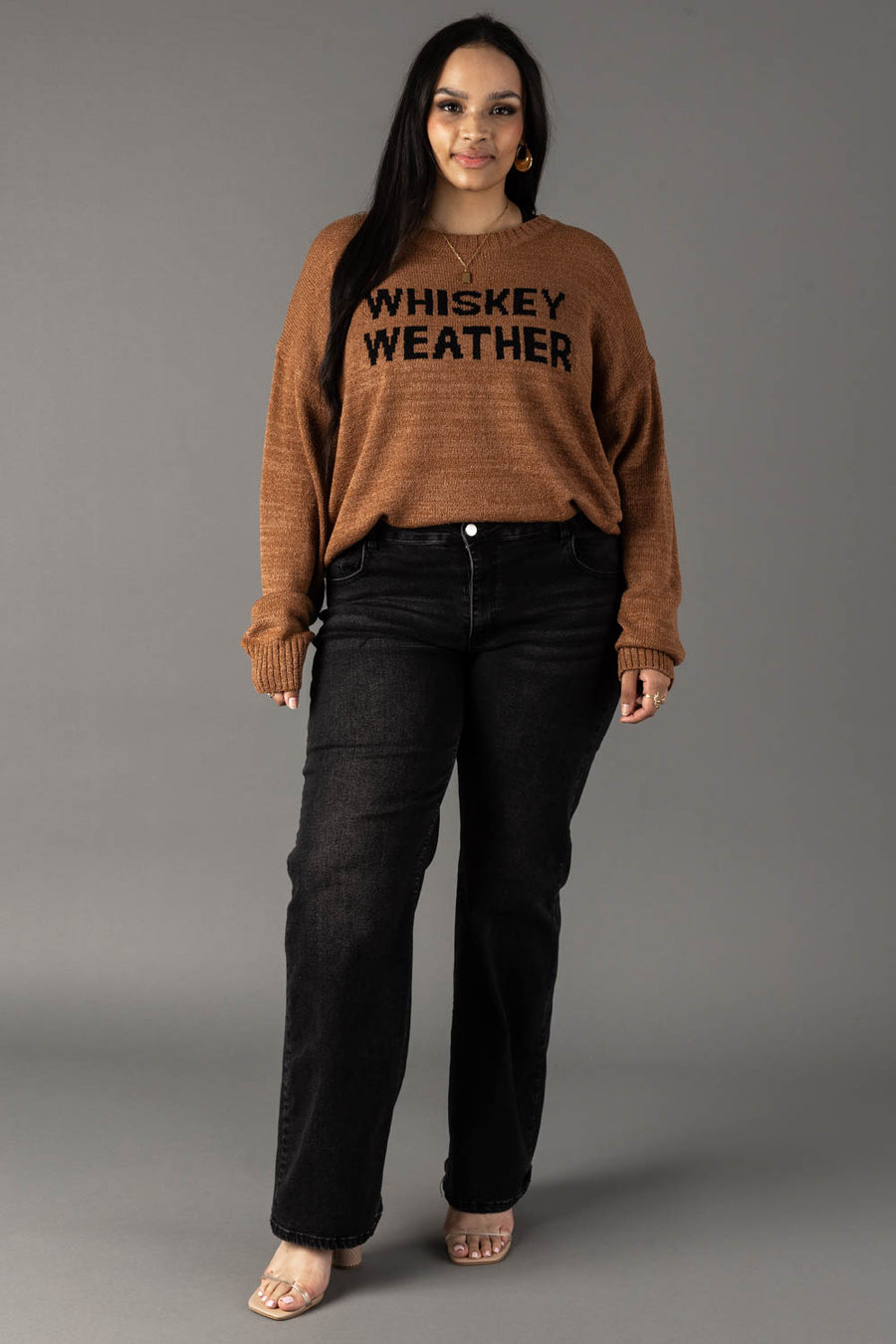 A woman with long dark hair stands against a gray background, wearing the Panache Apparel Collection's Brown Whiskey Weather Lightweight Sweater, paired with black jeans and clear-heeled sandals, smiling softly.