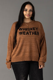 A woman with long dark hair models the Panache Apparel Collection's Brown Whiskey Weather Lightweight Sweater, featuring black lettering, paired with dark jeans against a gray background.
