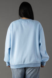 A person with long dark hair, wearing the Panache Apparel Collection Light Blue Drink On A Boat Sweatshirt and light blue jeans, stands facing away from the camera against a plain gray background.
