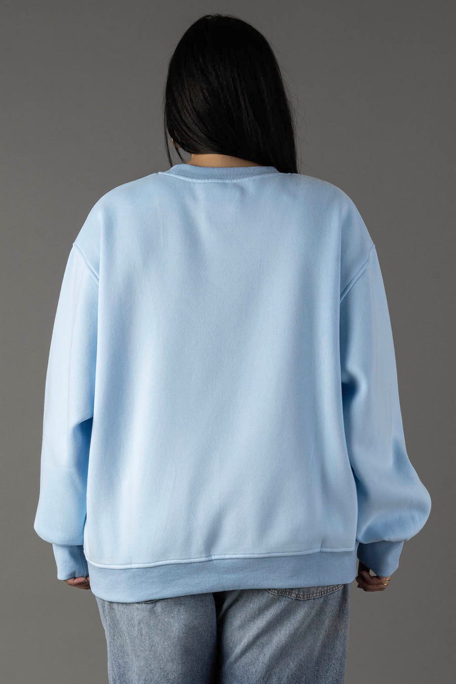 A person with long dark hair, wearing the Panache Apparel Collection Light Blue Drink On A Boat Sweatshirt and light blue jeans, stands facing away from the camera against a plain gray background.