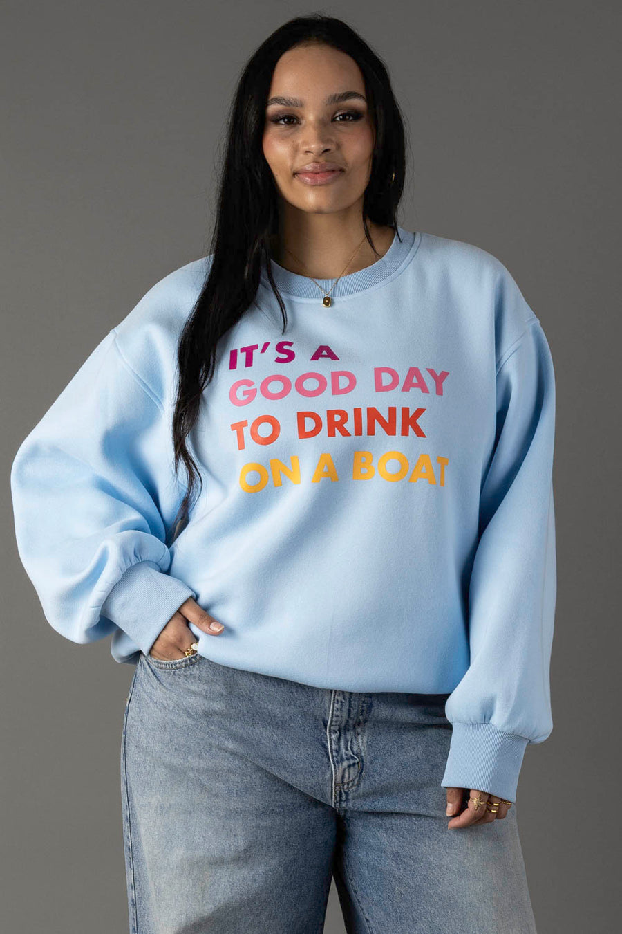 A woman wears the Panache Apparel Collection Light Blue Drink On A Boat Sweatshirt, featuring colorful "ITS A GOOD DAY TO DRINK ON A BOAT" text, as she stands against a gray background with one hand in her jeans pocket and a slight smile.