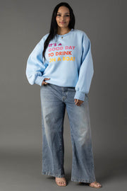 A woman stands against a gray background wearing the Panache Apparel Collection Light Blue Drink On A Boat Sweatshirt, featuring colorful text, styled with wide-leg jeans and open-toed heels.