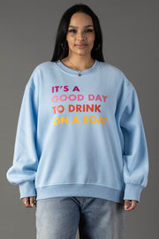 A woman in the Panache Apparel Collection Light Blue Drink On A Boat Sweatshirt with colorful “IT’S A GOOD DAY TO DRINK ON A BOAT” text stands against a gray background, wearing jeans and her long dark hair down.