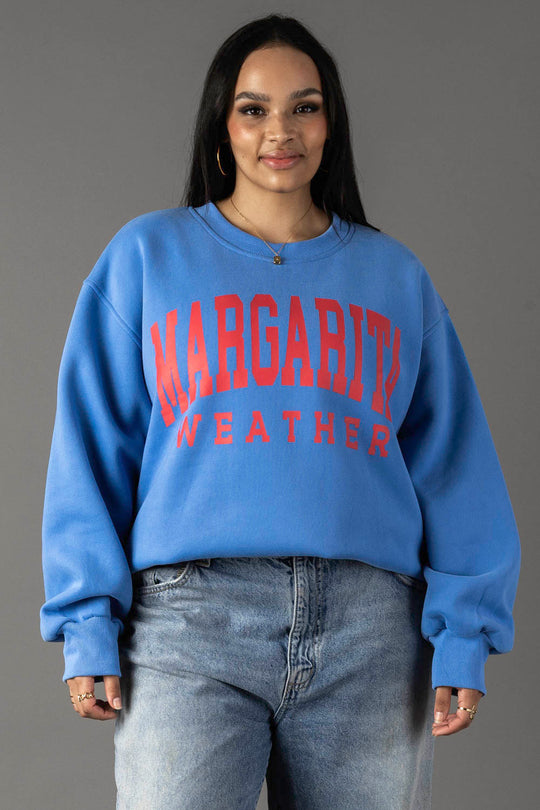 A woman with long dark hair wears the Blue Margarita Weather Sweatshirt from Panache Apparel Collection and light blue jeans, standing against a plain gray background.