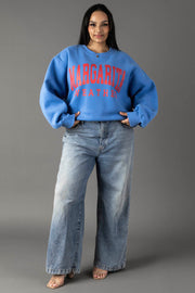 A woman wears the Blue Margarita Weather Sweatshirt by Panache Apparel Collection and wide-leg light blue jeans, standing against a gray background and smiling with her hands in her pockets.