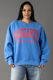A woman wears the Blue Margarita Weather Sweatshirt by Panache Apparel Collection, styled with light blue jeans. She stands against a plain gray background, highlighting the cozy oversized fleece crewneck.
