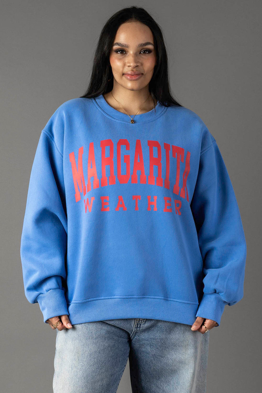 A woman wears the Blue Margarita Weather Sweatshirt by Panache Apparel Collection, styled with light blue jeans. She stands against a plain gray background, highlighting the cozy oversized fleece crewneck.