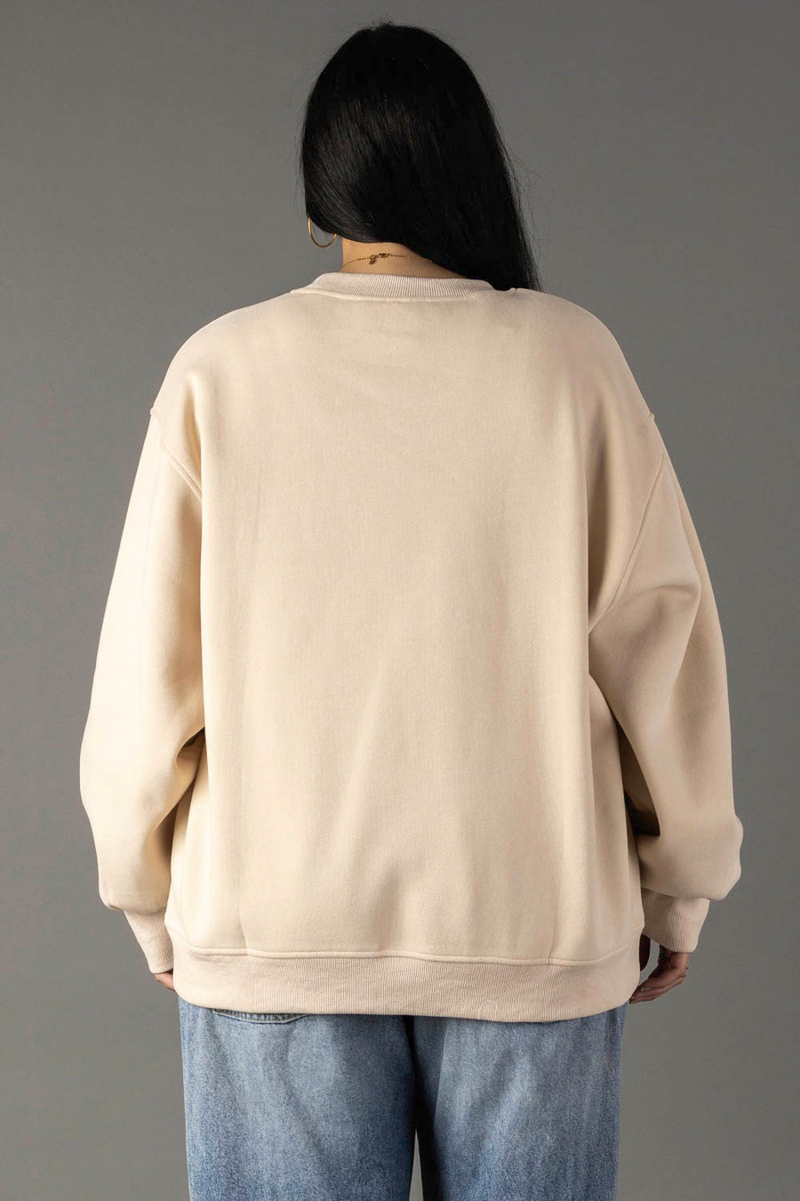 A person with long dark hair, shown from the back, wears a Tan Coffee Weather Sweatshirt by Panache Apparel Collection and light blue jeans against a plain gray background.