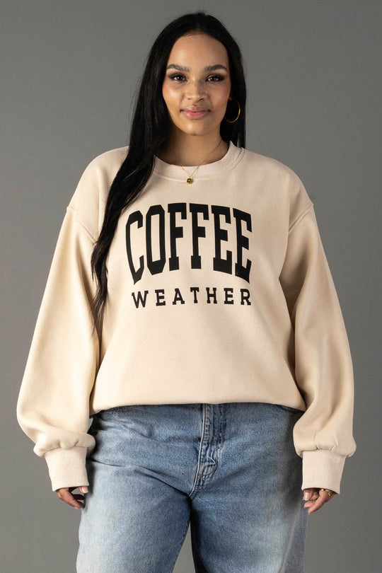 A woman with long dark hair wears hoop earrings and a Tan Coffee Weather Sweatshirt from Panache Apparel Collection, paired with blue jeans. She smiles slightly while standing against a plain gray background.