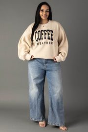 A woman smiles against a gray background, wearing the Panache Apparel Collection's Tan Coffee Weather Sweatshirt with bold black lettering, paired with light blue wide-leg jeans and open-toed heels.