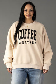 A woman with long dark hair wears a Tan Coffee Weather Sweatshirt from Panache Apparel Collection and light blue jeans, standing against a plain gray background.